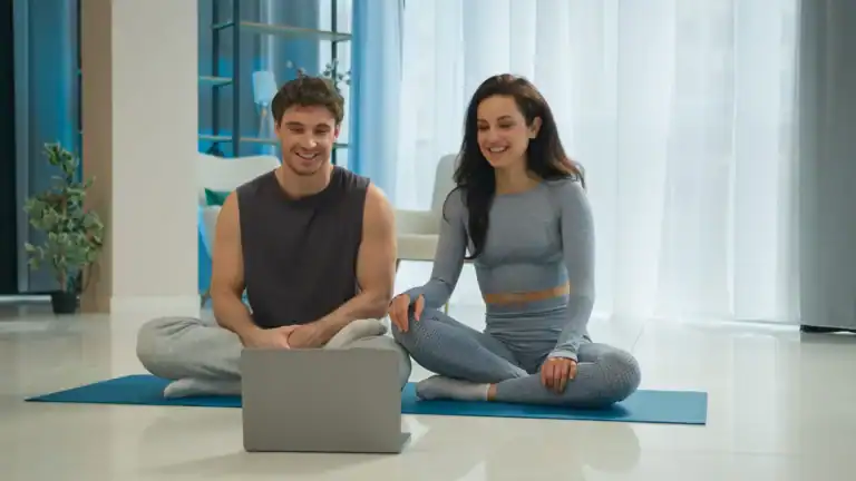 Ein junges Paar sitzt in sportlicher Kleidung auf Yogamatten vor einem Laptop und lächelt – symbolisiert gemeinsames Üben und die Vorteile von Yoga zu Hause in entspannter, moderner Umgebung.