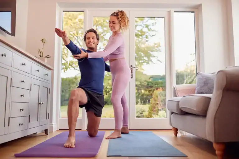 Yoga zu Hause: Eine Frau hilft einem Mann bei einer Yoga-Übung im Wohnzimmer – eine einfache und praktische Antwort auf die Frage: Was ist Yoga einfach erklärt.