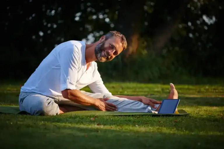 Ein Mann praktiziert Yoga im Freien und folgt einem Online-Kurs auf seinem Tablet. Diese Szene zeigt, wie vielseitig Yoga Arten sein können – von entspannenden Dehnungen bis hin zu energetisierenden Übungen, sogar draußen in der Natur.
