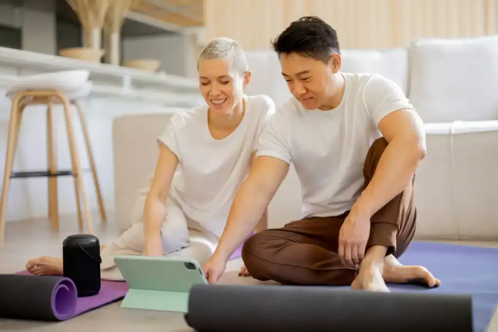Zwei Personen sitzen auf Yogamatten im Wohnzimmer und schauen gemeinsam ein Tablet an – ideal zur Teilnahme an Yoga Kursen online und zum Yoga online lernen von zu Hause aus.
