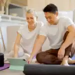 Zwei Personen sitzen auf Yogamatten im Wohnzimmer und schauen gemeinsam ein Tablet an – ideal zur Teilnahme an Yoga Kursen online und zum Yoga online lernen von zu Hause aus.