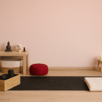 Ein liebevoll eingerichteter Raum für Yoga zu Hause: Auf einer schwarzen Yogamatte mit Blumenmuster liegt eine gefaltete Decke bereit. Daneben stehen ein geflochtener Korb mit Yoga-Zubehör und ein rotes Meditationskissen. Ein kleiner Tisch mit Buddha-Figur, Duftlampe und ätherischen Ölen unterstreicht die ruhige Atmosphäre – ideal für Yoga daheim.