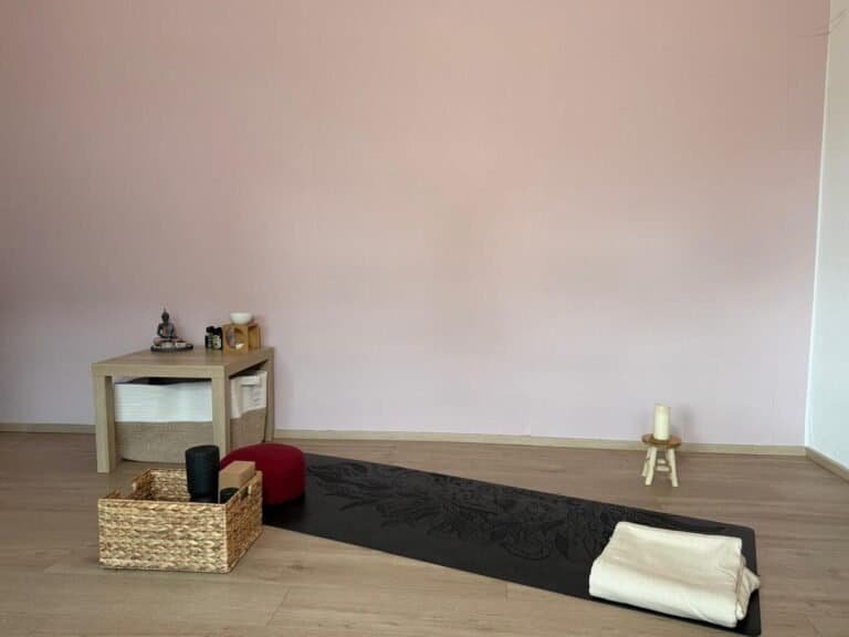 Ein liebevoll eingerichteter Raum für Yoga zu Hause: Auf einer schwarzen Yogamatte mit Blumenmuster liegt eine gefaltete Decke bereit. Daneben stehen ein geflochtener Korb mit Yoga-Zubehör und ein rotes Meditationskissen. Ein kleiner Tisch mit Buddha-Figur, Duftlampe und ätherischen Ölen unterstreicht die ruhige Atmosphäre – ideal für Yoga daheim.