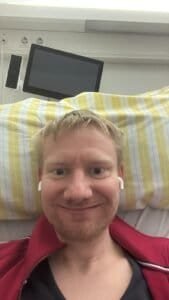 David Train im Krankenhausbett, einen Tag vor seiner Herzoperation, lächelnd mit Ohrhörern – ein Moment voller Mut und Zuversicht.