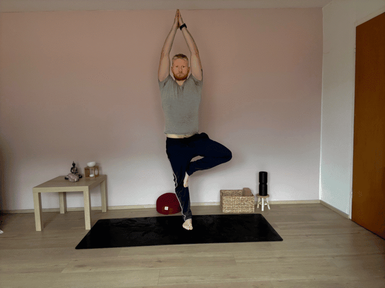 Mann übt Vrikshasana, die Baumhaltung im Yoga. Ein Fuß steht fest auf der Matte, der andere liegt am Oberschenkel, Hände über dem Kopf gefaltet. Diese Yoga Übung fördert Gleichgewicht, Konzentration und innere Ruhe.