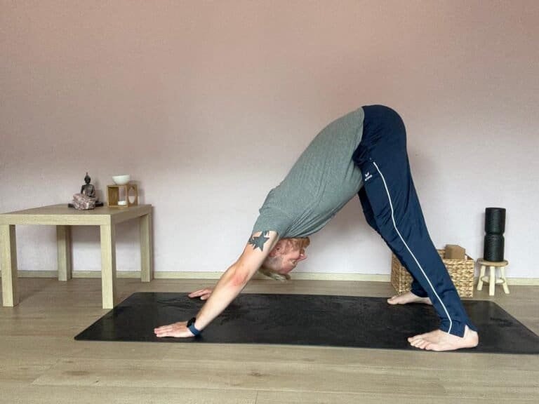 Mann übt Adho Mukha Shvanasana, den herabschauenden Hund im Yoga. Hände und Füße fest auf der Matte, Gesäß nach oben gestreckt, Rücken gerade. Aufnahme im Yogaraum mit Tisch, Buddha-Figur und Yoga-Accessoires im Hintergrund.