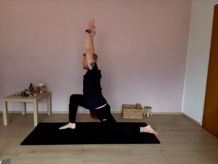Mann übt Yoga am Morgen in der Pose Anjaneyasana (Low Lunge) auf einer Matte, mit gestreckten Armen nach oben, zur Dehnung und Hüftöffnung im Wohnzimmer.