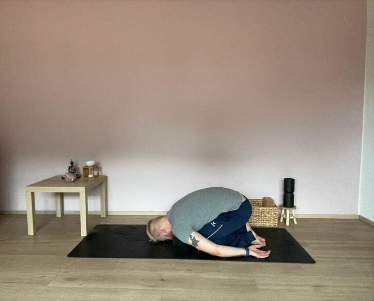 Mann in Balasana, der Kindeshaltung im Yoga. Stirn auf der Matte, Arme nach hinten neben dem Körper abgelegt, Schultern entspannt. Aufnahme in einem ruhigen Yogaraum mit Buddha-Figur und Yoga-Accessoires im Hintergrund.