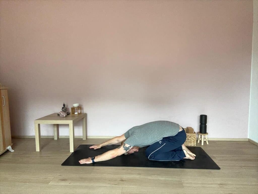 Mann übt Balasana, die Kindeshaltung im Yoga, mit nach vorne ausgestreckten Armen. Stirn liegt auf der Matte, Rücken entspannt, Gesäß sinkt zu den Fersen. Im Hintergrund ein kleiner Yogaraum mit Tisch, Buddha-Figur und Meditationszubehör.