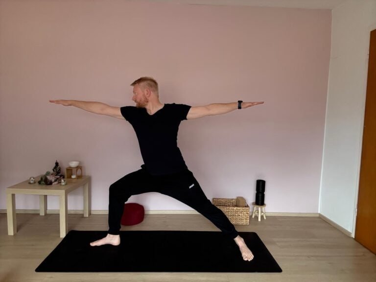 Mann übt Virabhadrasana II, den Krieger 2 im Yoga. Vorderes Bein ist im rechten Winkel gebeugt, hinteres Bein gestreckt, Arme parallel zum Boden ausgestreckt. Diese Yoga Übung stärkt Beine und Rumpf, verbessert die Balance und fördert innere Stabilität.