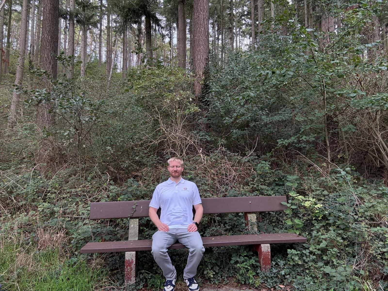 Mann sitzt entspannt auf einer Bank im Wald, umgeben von Bäumen und Grün – ein Moment der Ruhe in der Natur, ideal um das Nervensystem zu beruhigen