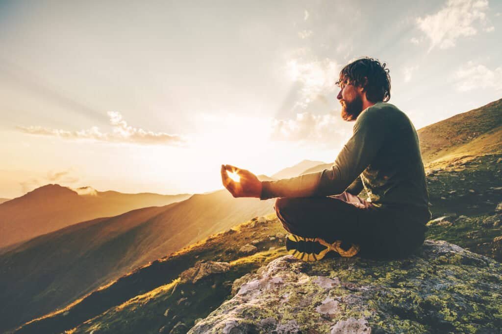 Meditation Erfahrungen in der Natur – Mann sitzt im Schneidersitz auf einem Berg und meditiert bei Sonnenuntergang, um Ruhe und innere Balance zu finden.