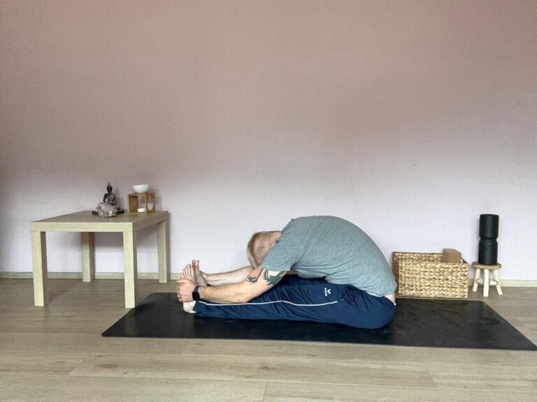 Mann übt Paschimottanasana, die sitzende Vorbeuge im Yoga. Beine sind lang nach vorne ausgestreckt, Hände umfassen die Füße, Oberkörper nach vorne über die Beine geneigt. Aufnahme in einem ruhigen Yogaraum mit Tisch, Buddha-Figur und Meditationszubehör.