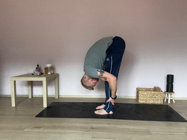 Mann übt Uttanasana, die stehende Vorbeuge im Yoga. Oberkörper hängt entspannt über den gestreckten Beinen, Hände umfassen die Unterschenkel. Diese Yoga Übung dehnt die Beinrückseiten, entspannt den Rücken und beruhigt das Nervensystem.