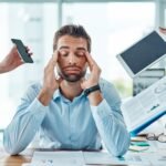 Gestresster Mann im Büro mit geschlossenen Augen, der sich die Schläfen reibt, während ihm gleichzeitig ein Smartphone, eine Uhr, ein Tablet und Unterlagen gezeigt werden – symbolische Darstellung für Überforderung und Dauerstress Symptome im Arbeitsalltag.