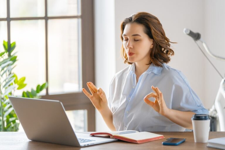 Frau sitzt entspannt am Schreibtisch vor dem Laptop und übt eine Atemtechnik zur Beruhigung des Vagusnervs. Mit geschlossenen Augen und ruhiger Haltung praktiziert sie bewusstes Atmen, um Stress abzubauen und das Nervensystem zu entspannen.