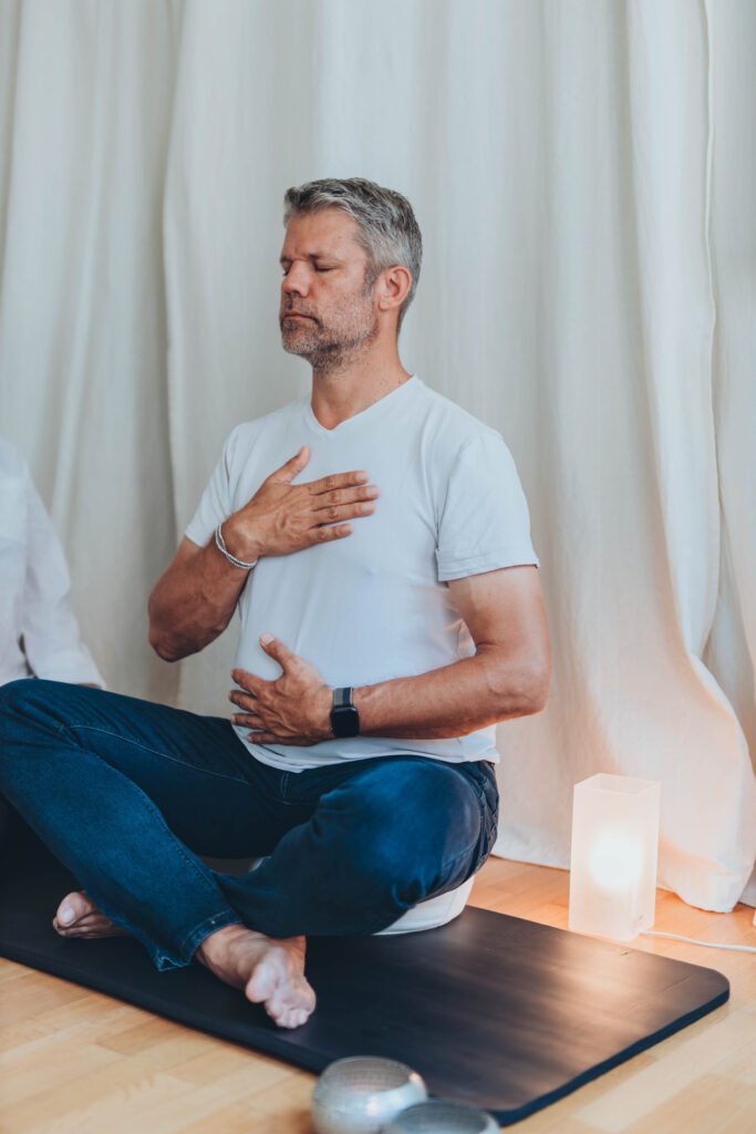 Mann sitzt im Schneidersitz auf einer Yogamatte, Augen geschlossen, eine Hand auf Brust und eine auf Bauch – Atem Meditation und kohärente Atmung zur Beruhigung des Nervensystems und Förderung innerer Balance.