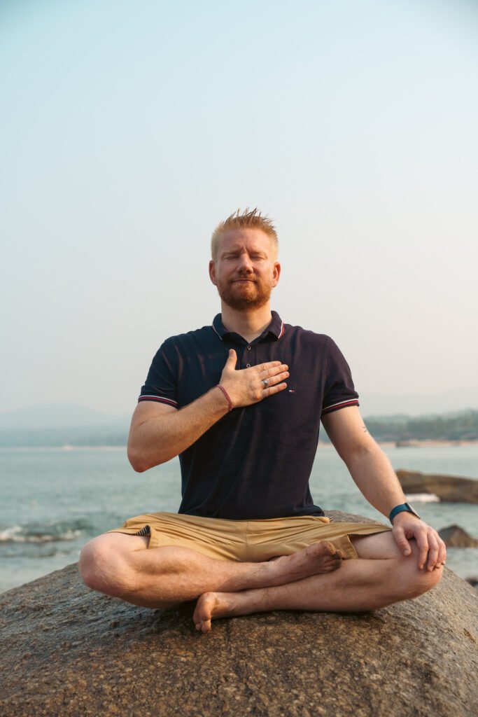 Mann sitzt meditierend im Schneidersitz auf einem Felsen am Meer und praktiziert Herzkohärenz Atmung und Herzkohärenz Meditation zur Beruhigung des Nervensystems.