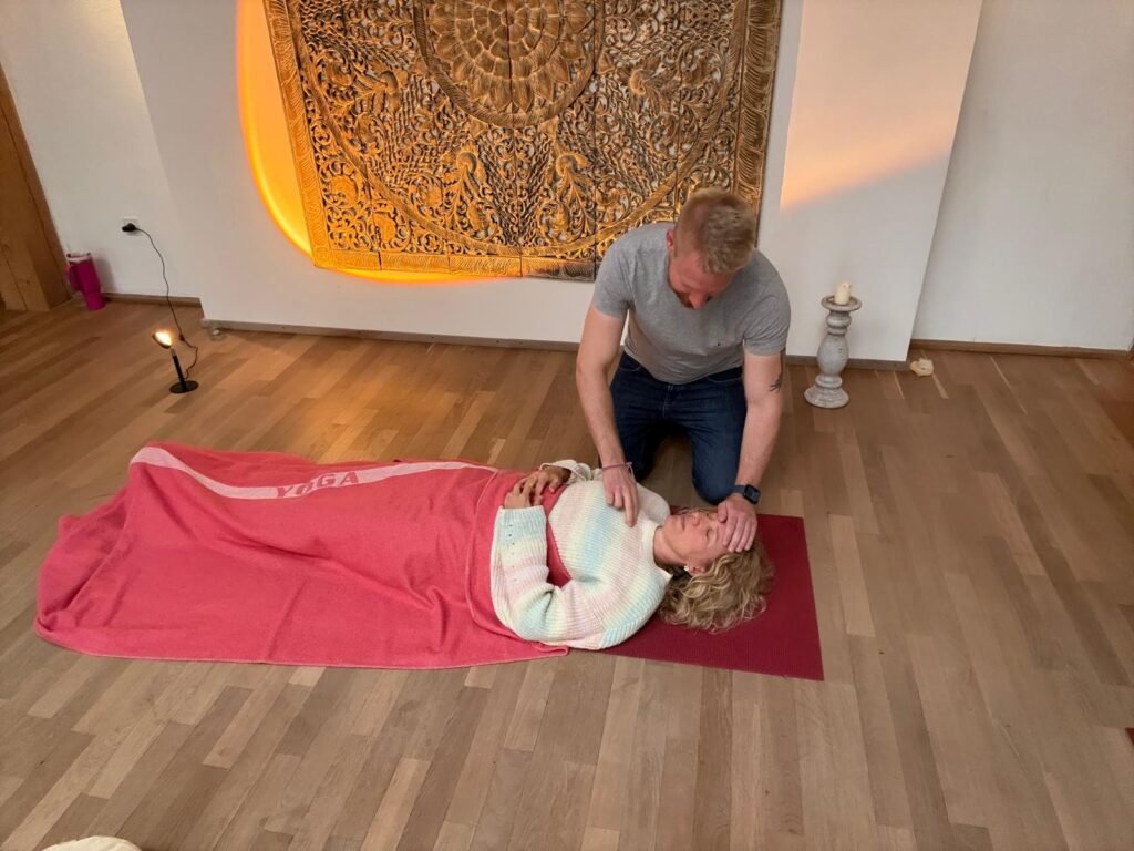 Breathwork Coach David Train begleitet eine Teilnehmerin während einer Transformative Breathwork Session – sie liegt entspannt auf einer Yogamatte, zugedeckt mit einer Decke, während David achtsam Hände auf Bauch und Stirn legt und sie durch den Atemprozess führt.
