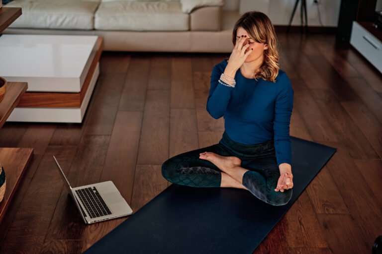 Eine Frau sitzt im Schneidersitz auf einer dunklen Yogamatte und praktiziert Nadi Shodhana (Wechselatmung), eine klassische Pranayama-Atemübung aus dem Breathwork-Bereich. Sie hält eine Hand vor ihr Gesicht, um die Nasenlöcher abzuwechseln. Vor ihr steht ein aufgeklappter Laptop, im Hintergrund ein helles Wohnzimmer mit Holzboden.