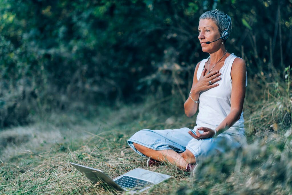 Frau mit kurzem grauem Haar sitzt im Schneidersitz auf einer Wiese in der Natur, trägt ein Headset und hält eine Hand auf ihr Herz während einer transformative breathwork session online – vor ihr steht ein aufgeklappter