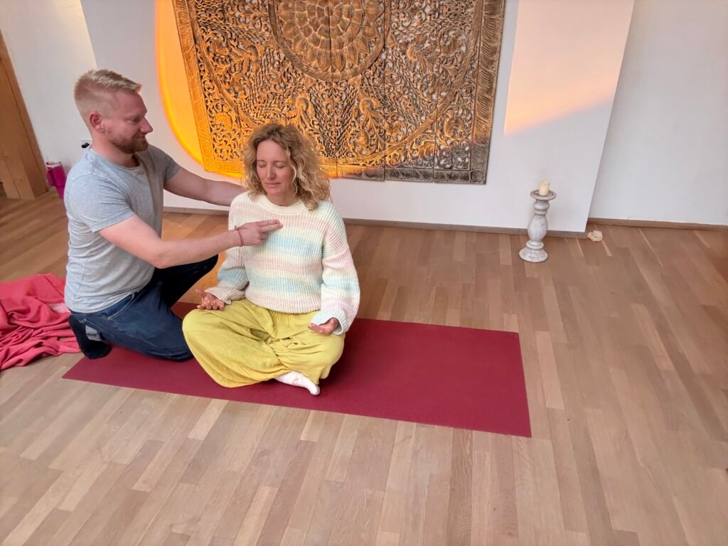Breathwork Coach David Train begleitet eine Teilnehmerin während einer Transformative Breathwork Session – eine intensive Psychedelic Breath Erfahrung auf einer roten Yogamatte. Er sitzt achtsam hinter ihr und führt sie durch den Atemprozess. Im Hintergrund ein warmes Ambiente mit einem kunstvollen Holzrelief und einer Kerze.