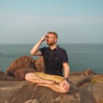 Person praktiziert Atemübungen zur Entspannung am Meer und macht in meditativer Sitzhaltung Atemübungen gegen Stress und Angst.