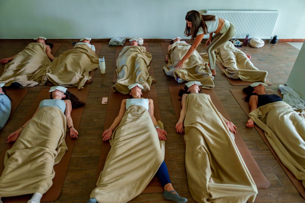 Eine transformative Breathwork-Gruppensession: Neun Teilnehmende liegen in Reihen auf Yogamatten, eingehüllt in warme beige Decken und mit weißen Augenmasken, um vollständig in ihre Breathwork-Erfahrung eintauchen zu können. Eine Facilitatorin beugt sich fürsorglich zu einer Teilnehmerin herunter und begleitet sie achtsam durch den Prozess. Der helle, schlichte Raum schafft eine ruhige und sichere Atmosphäre für die innere Reise.