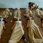 Eine transformative Breathwork-Gruppensession: Neun Teilnehmende liegen in Reihen auf Yogamatten, eingehüllt in warme beige Decken und mit weißen Augenmasken, um vollständig in ihre Breathwork-Erfahrung eintauchen zu können. Eine Facilitatorin beugt sich fürsorglich zu einer Teilnehmerin herunter und begleitet sie achtsam durch den Prozess. Der helle, schlichte Raum schafft eine ruhige und sichere Atmosphäre für die innere Reise.