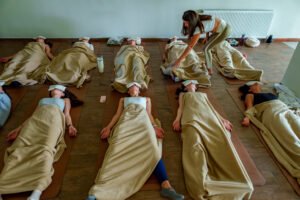 Eine transformative Breathwork-Gruppensession: Neun Teilnehmende liegen in Reihen auf Yogamatten, eingehüllt in warme beige Decken und mit weißen Augenmasken, um vollständig in ihre Breathwork-Erfahrung eintauchen zu können. Eine Facilitatorin beugt sich fürsorglich zu einer Teilnehmerin herunter und begleitet sie achtsam durch den Prozess. Der helle, schlichte Raum schafft eine ruhige und sichere Atmosphäre für die innere Reise.