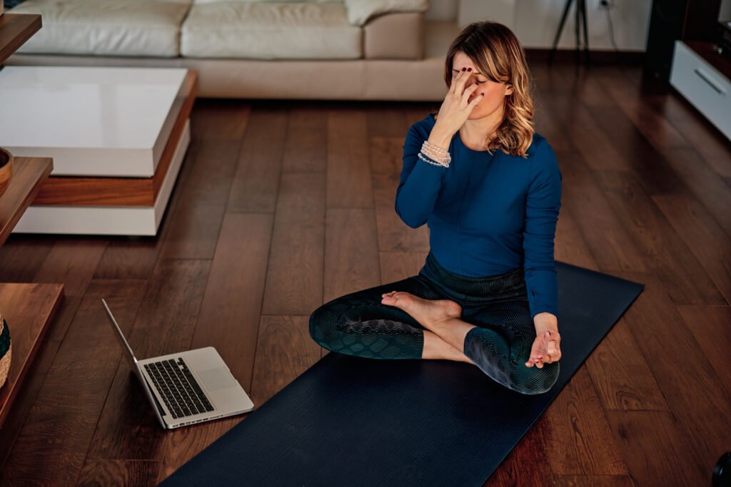 Woman lying relaxed on a yoga mat and practicing conscious breathing with closed eyes while connected via laptop – scene from a transformative breathwork session online for emotional regulation and