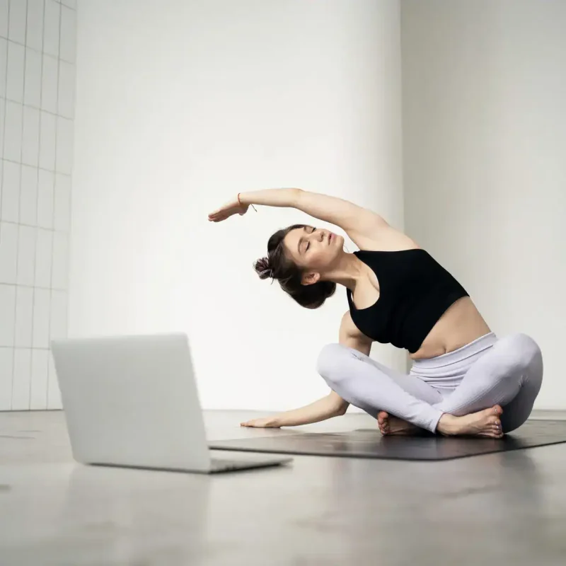Eine junge Frau praktiziert zuhause Yoga mit einem Laptop, während sie eine seitliche Dehnung im Schneidersitz ausführt. Diese Szene steht exemplarisch für moderne Arten von Yoga, die sich flexibel in den Alltag integrieren lassen – besonders durch Online-Kurse.