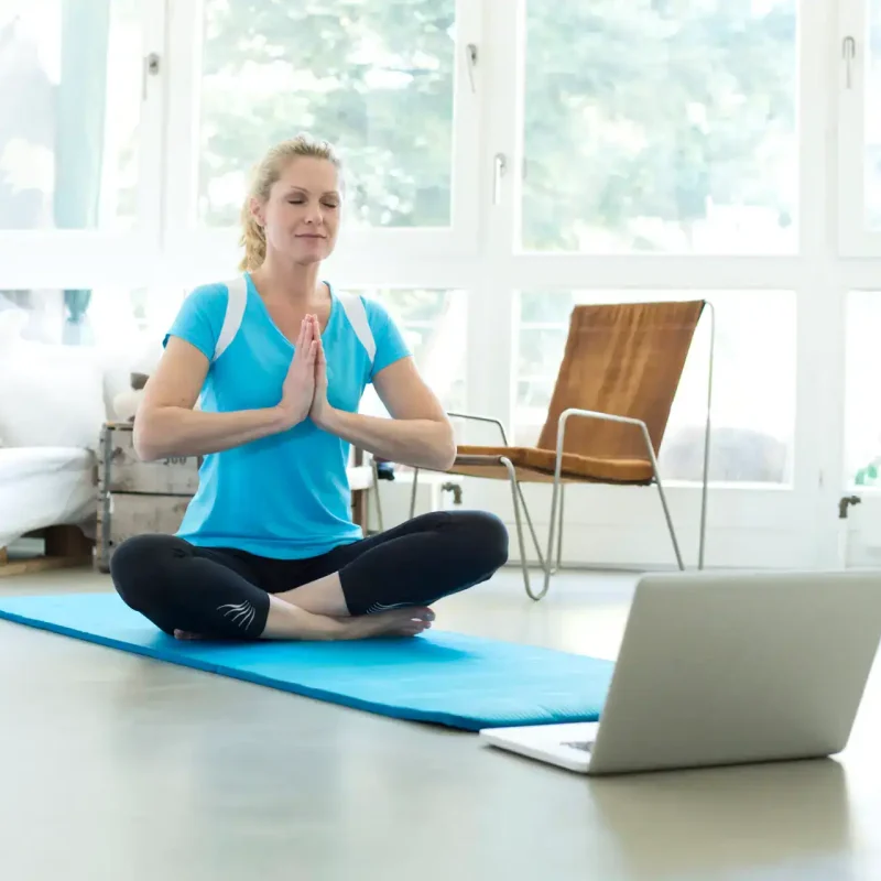 Frau sitzt in Meditationshaltung auf einer Yogamatte vor dem Laptop – ein Moment der Achtsamkeit mit einem der besten Yoga Kurse online für Zuhause.